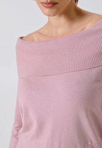 Maglione leggero rosa chiaro senza spalline in un morbido tessuto texturizzato con collo a coste. Dettaglio di branding discreto in basso a destra.