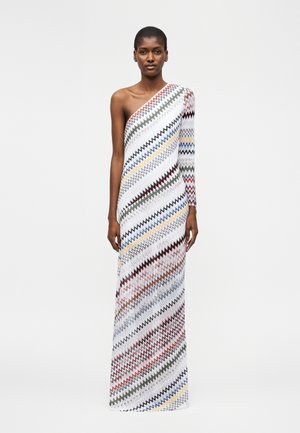 Model trägt ein bodenlanges, einseitiges Kleid mit diagonalen, mehrfarbigen Zickzackmustern auf weißem Hintergrund.