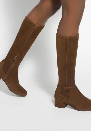 Bottes hautes en daim marron avec petits talons carrés portées sur des jambes sur un fond clair uni.