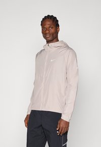 Lys beige Nike hættejakke med fuld lynlås, minimalistisk design og et lille hvidt logo på brystet. Båret med mørke shorts.