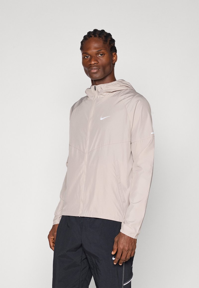 Lys beige Nike hættejakke med fuld lynlås, minimalistisk design og et lille hvidt logo på brystet. Båret med mørke shorts.