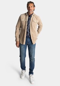Beige corduroy shirt met twee borstzakken, gedragen over een donkerblauw overhemd, gecombineerd met blauwe spijkerbroek en witte sneakers.
