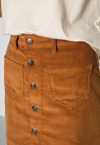 Bruine corduroy rok met verticale ribbelstructuur, voorzien van zilveren knopen aan de voorkant, twee zakken en een aansluitende taille.