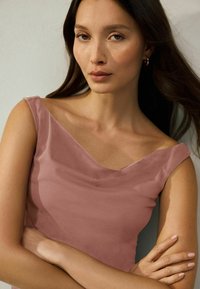 Topy s krátkym rukávom, hlbokým V-vystřihem v mäkkej ružovej látke. Hladká textúra s jemným leskom. Modelka s prekríženými rukami a neutrálnym pozadím.