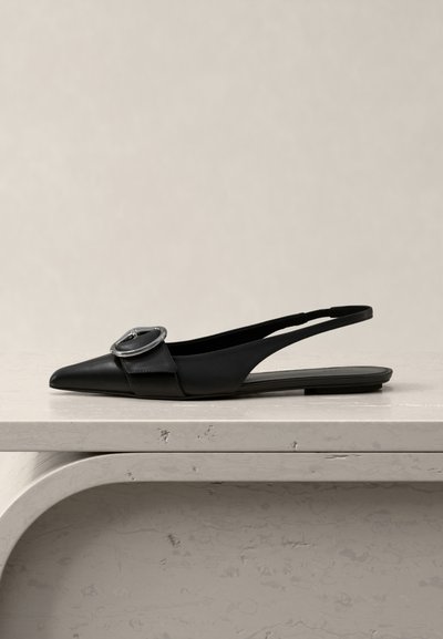 Μαύρο, μυτερό, ίσιο πέδιλο με λουράκι στο πίσω μέρος και μεγάλη ασημένια αγκράφα, τοποθετημένο σε μπεζ μαρμάρινη επιφάνεια σε ουδέτερο φόντο.