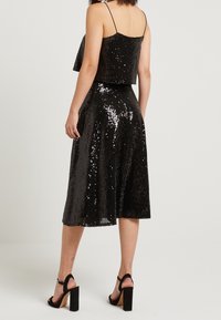 Black sequined crop top paired with a knee-length sequined skirt. Features adjustable straps and a flared silhouette. Black block-heeled sandals.