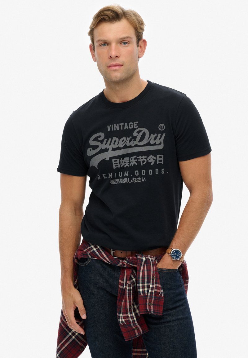 Camiseta de algodón negra con texto gráfico en gris en el frente, combinada con jeans oscuros y una camisa de cuadros roja atada a la cintura. Reloj marrón en la muñeca.