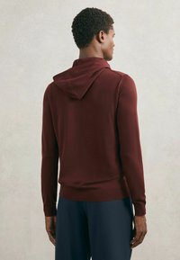 Bordó pletená mikina s kulatou kapucí, přiléhavými rukávy a žebrovaným lemem. Nošena s volnými tmavě modrými kalhotami.