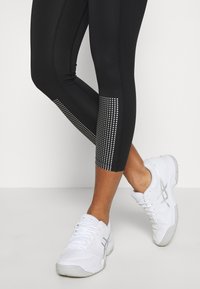 Czarne legginsy sportowe z teksturowanym, refleksyjnym wzorem na dolnej części nogawki. Połączone z białymi butami sportowymi z jasnoszarymi akcentami.