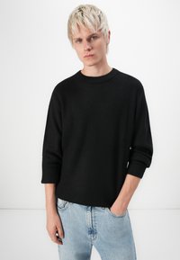 Pull en texture noire avec un col rond et des manches trois-quarts, associé à un jean en denim bleu clair. Le tissu semble épais et confortable.