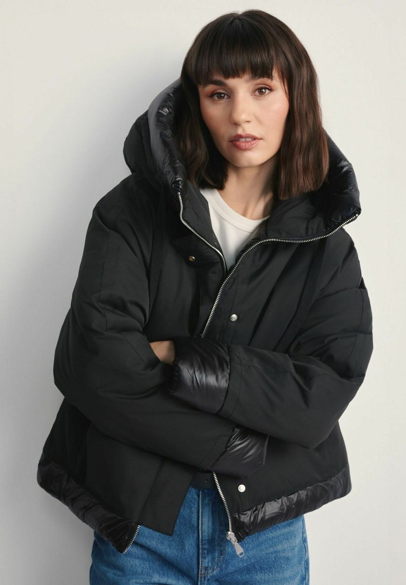 Czarna kurtka puchowa z dużym kapturem, błyszczące wykończenie na kapturze i mankietach, zamek błyskawiczny i krótszy fason. Połączona z niebieskimi dżinsami.