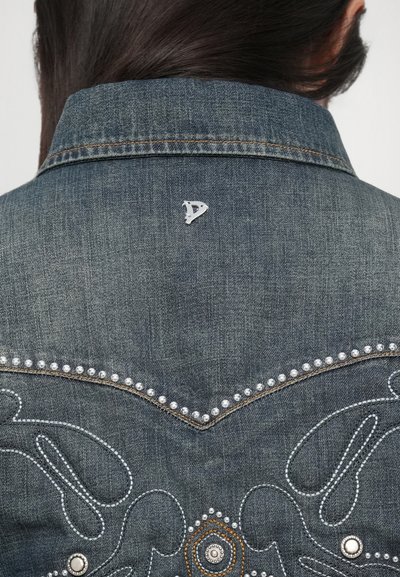 Denim jack met een donkere blauwe kleur, decoratieve zilveren studs langs de naden en een geborduurd ontwerp op de achterkant met een logo-pin.