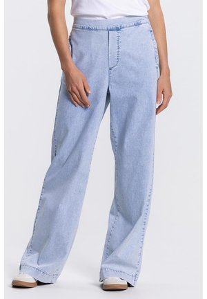 Persoon draagt lichtblauwe wijd uitlopende spijkerbroek, witte sneakers en een wit shirt, staand tegen een effen achtergrond.