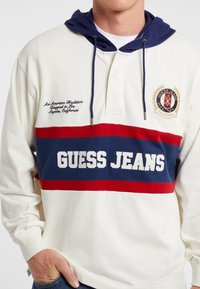 Langsleeve hoodie met een crèmekleurig lichaam, marineblauwe capuchon en gedurfde rode en blauwe strepen. Bevat het "GUESS JEANS" logo en geborduurde accenten.