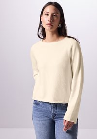 Maglione lungo a maniche concolore crema, con scollatura rotonda, realizzato in tessuto morbido, caratterizzato da una vestibilità rilassata e polsini a coste. Abbinato a jeans blu.