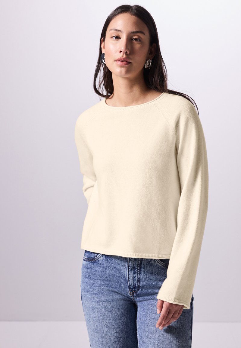 Maglione lungo a maniche concolore crema, con scollatura rotonda, realizzato in tessuto morbido, caratterizzato da una vestibilità rilassata e polsini a coste. Abbinato a jeans blu.
