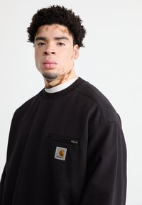Juodas džemperis su užtrauktuku ir mažu Carhartt logotipo ženkliuku. Turi apvalią kaklo iškirptę ir laisvą siluetą, pagamintas iš minkštos medžiagos.