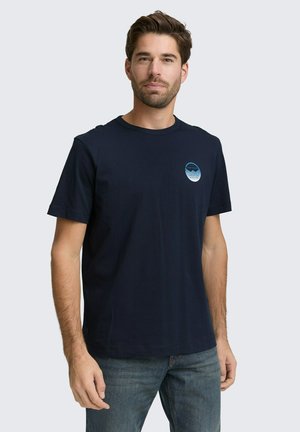 Man draagt een marineblauw T-shirt met korte mouwen en een kleine ronde logo op de linkerborst, gecombineerd met een blauwe spijkerbroek, staand tegen een effen achtergrond.
