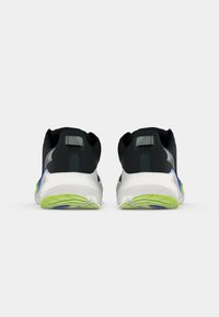 Baskets de sport noires avec semelles blanches, accents verts et une section arrière texturée avec une bande réfléchissante. Design minimaliste.