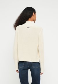 Pulover tricotat de culoare crem, cu guler înalt și manșete drepte, având o etichetă mică cu logo în partea din spate. Purtați cu blugi albastru închis.