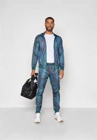 Chándal en degradado azul-verde con detalles blancos, chaqueta con cremallera, pantalones con cintura elástica y una bolsa de lona negra. Zapatillas blancas.