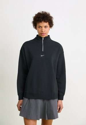 Sweatshirt zippé noir avec col haut, arborant un logo Nike blanc sur la poitrine. Matière douce, coupe décontractée, associé à une jupe plissée grise.