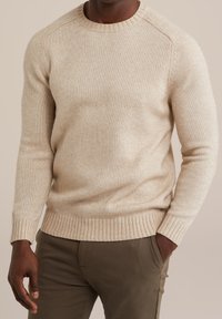 Béžový pletený svetr s žebrovaným kulatým výstřihem a manžetami, s texturovaným povrchem. Nosit s olivově zelenými kalhotami.