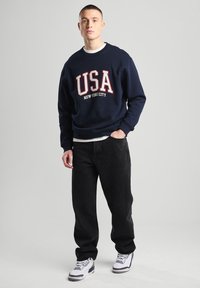 Navy sweatshirt with "USA NEW YORK CITY" in red and white. Paired with black pants and white sneakers. Simple design, casual style.