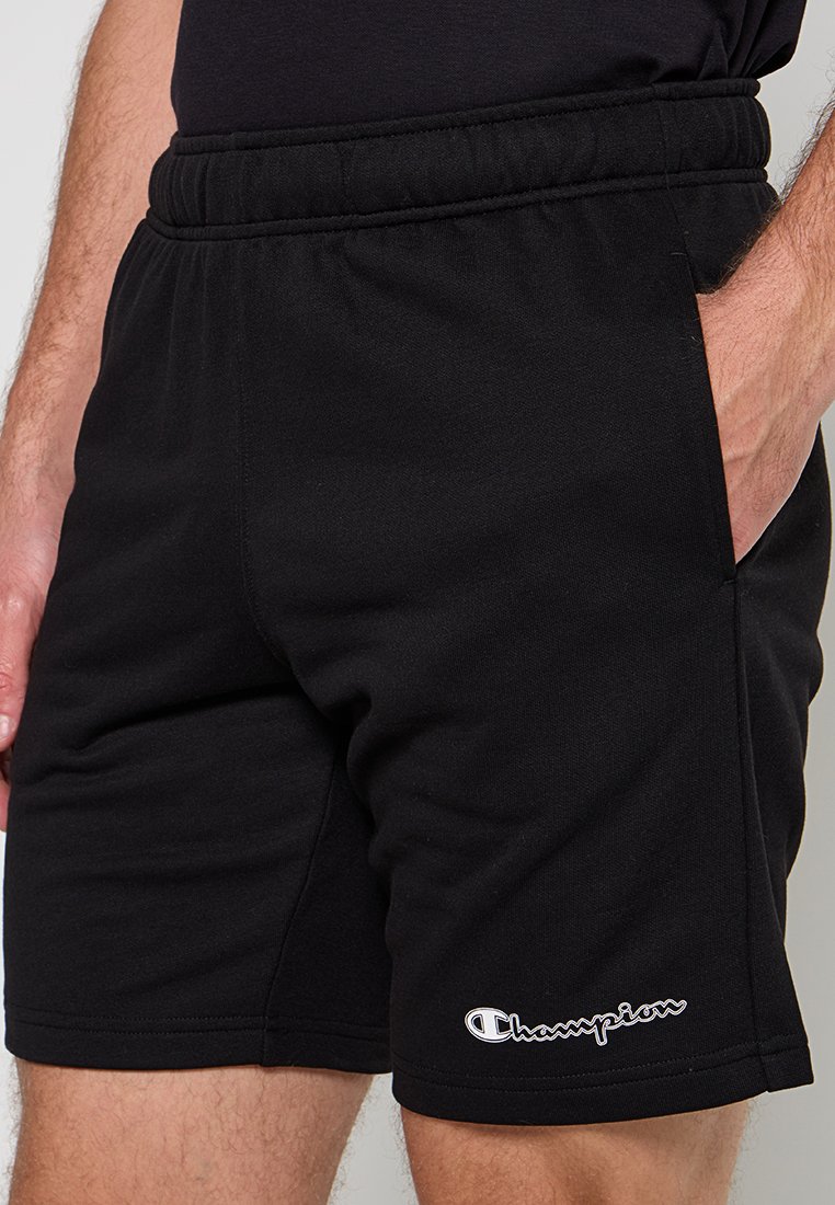 Hombre con pantalones cortos negros de Champion con cinturilla elástica y bolsillos laterales, mano descansando dentro del bolsillo derecho.