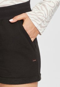 Schwarze Shorts mit einem gerippten Bund, Seitentaschen und aufgerolltem Saum. Auffälliges Detail ist ein kleines Markenetikett an der Seite.
