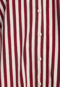 Balta krekls ar vertikālām sarkanām svītrām un baltajiem pogām, kas izvietotas gar centra priekšu.