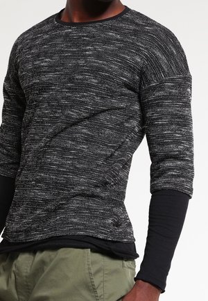 Mann, der geschichtete Kleidung trägt, mit einem schwarz-grau melierten Langarmpullover über einem schwarzen eng anliegenden Shirt und olivgrünen Hosen.