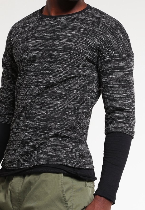 Homme portant des vêtements superposés avec un pull à manches longues noir et gris marbré sur une chemise noire ajustée et un pantalon vert olive.