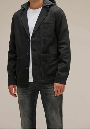 Mężczyzna ubrany w czarną kurtkę zapinaną na guziki z kapturem, pod którą ma białą koszulkę oraz wyblakłe czarne dżinsy, stoi na jednolitym tle.