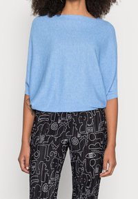 Pullover in maglia azzurro chiaro, morbido e ampio, con scollatura larga e spalle scese, abbinato a pantaloni neri con disegni astratti bianchi.