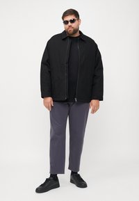 Zwarte ritssluitingjack over een zwart t-shirt, gecombineerd met grijze broeken en zwarte sneakers. De persoon draagt donkere zonnebrillen.
