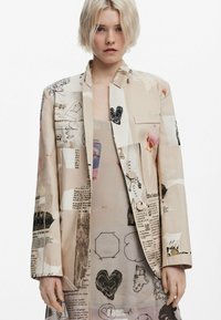 Beige blazer with mixed print, including text and graphics. Features a single chest pocket, notch lapels, and a relaxed fit.