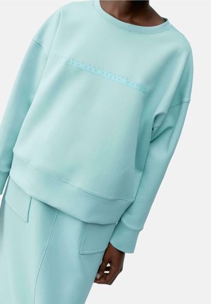 Person trägt hellblauen Pullover mit dezentem, geprägtem Schriftzug und passenden Rock mit großen Fronttaschen.