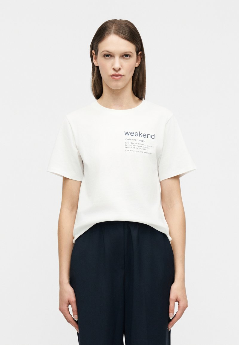 Γυναίκα με ίσια καστανά μαλλιά, ντυμένη με λευκό t-shirt με την επιγραφή "weekend" και μαύρο παντελόνι, στέκεται μπροστά σε ένα απλό φωτεινό φόντο.
