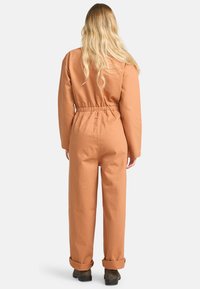 Oranje katoenen jumpsuit met lange mouwen, elastische taille en opgerolde mouwen. Heeft een eenvoudig ontwerp en een effen kleur in het hele kledingstuk.