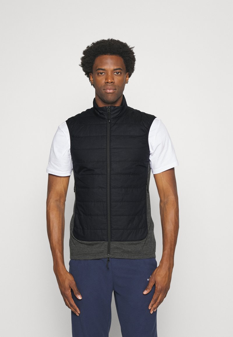 Schwarze gepolsterte Weste mit hohem Kragen und Reißverschluss, die über einem grauen Langarmshirt getragen wird. Das Modell zeigt einen neutralen Gesichtsausdruck.
