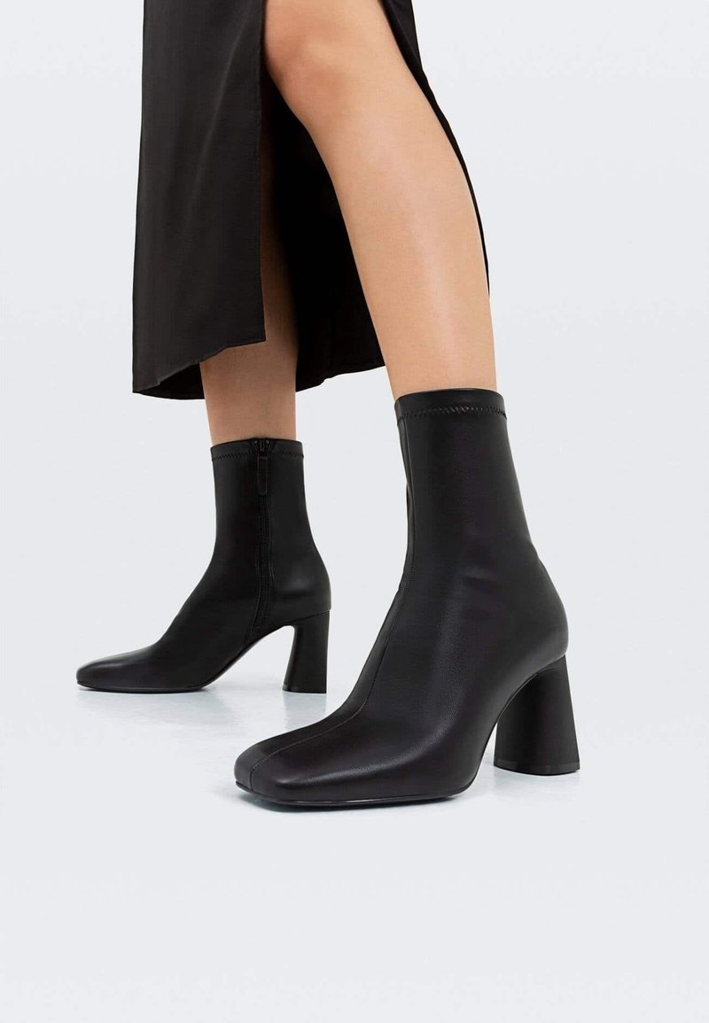 Jambes portant des bottes en cuir noir mi-mollet à talons blocs et fermetures éclair latérales, associées à une jupe noire avec une fente à l'avant.