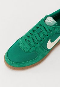 Baskets Nike vertes avec logo Swoosh blanc, lacets verts et semelle en gomme sur fond blanc.