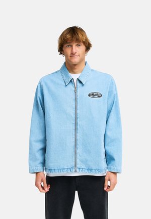 Jeune homme aux cheveux châtain clair portant une veste bleu clair zippée avec col et un petit logo sur la poitrine, debout devant un fond uni.