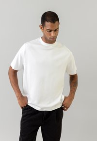 Tricou alb din bumbac, cu mâneci scurte și guler rotund. Purtați cu pantaloni închiși la culoare, expus pe un fundal gri deschis.