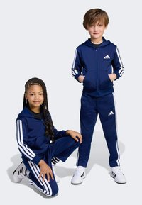 Námořnické teplákové soupravy s bílými pruhy, mikina na zip a elastický pás. Děti nosí tenisky se třemi pruhy a logovými akcenty.