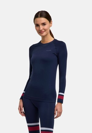 Kvinde med mørkt hår i en hestehale iført en tætsiddende marineblå bluse med lange ærmer og matchende leggings med røde og hvide striber.