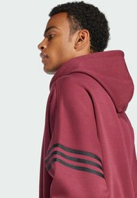Felpa color marrone con un design a strisce nere sulle maniche, realizzata in tessuto morbido. Presenta un cappuccio e una vestibilità comoda, con una texture liscia.
