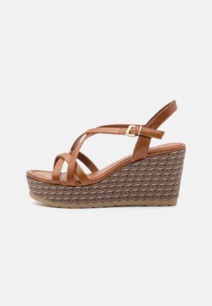 Brun kile sandal med krydsede stropper, ankelspænde og mønstret platformsål på en ensfarvet baggrund.