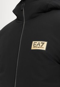 Jachetă neagră cu fermoar, dotată cu un patch rectangular EA7 Emporio Armani bej pe partea stângă a pieptului și cu un guler înalt.